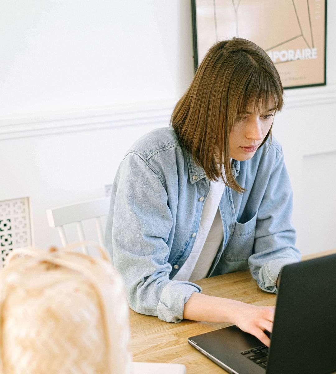 A woman in a denim shirt working on a laptop at a wooden desk in a bright room