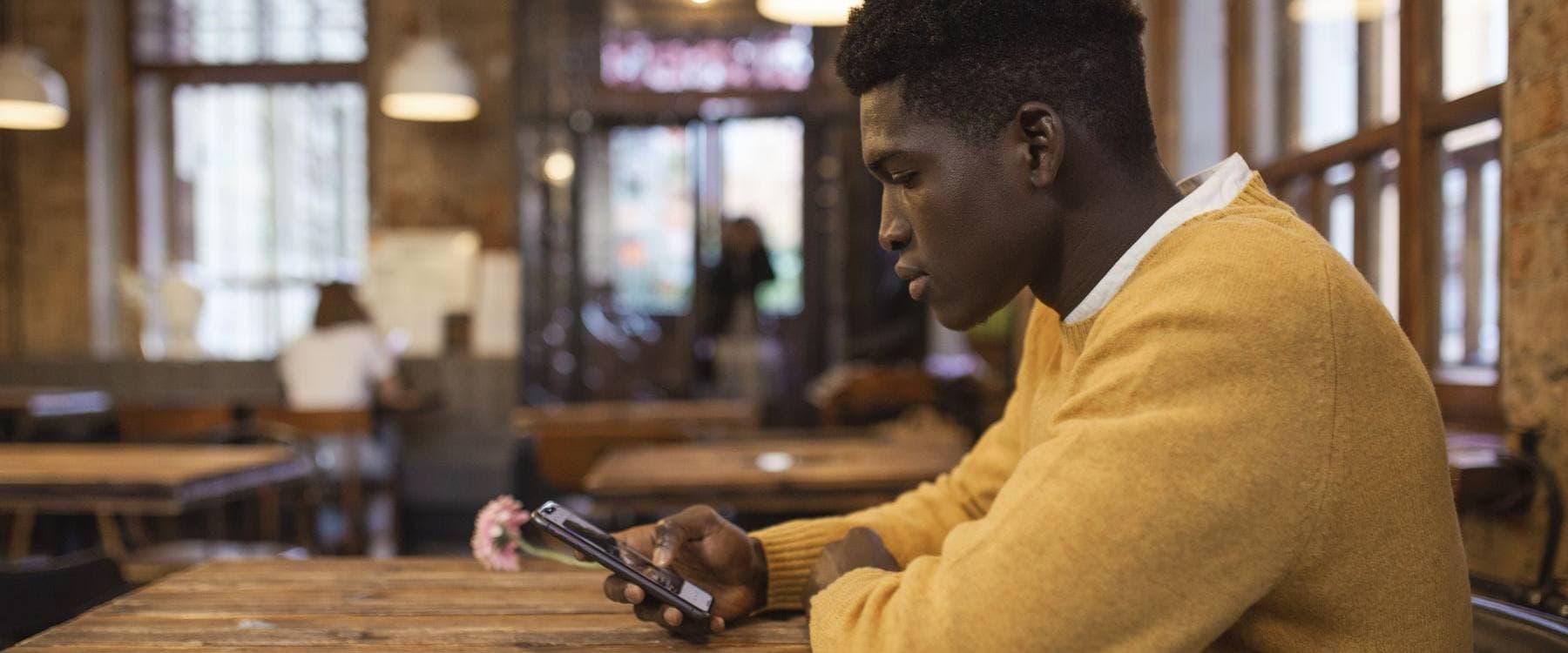 A man in a mustard sweater checks his phone at a wooden table in a cafe